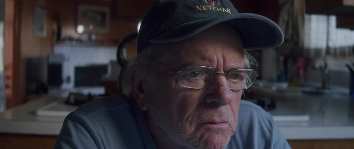 Portrait of an elderly man wearing a cap sitting down taking off his glasses