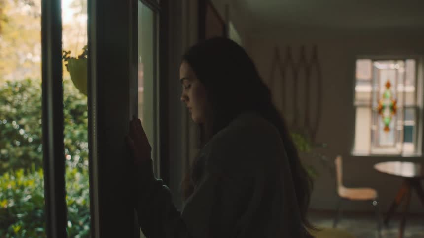 A woman reclines against a glass door in a sunlit living room gazing out longingly