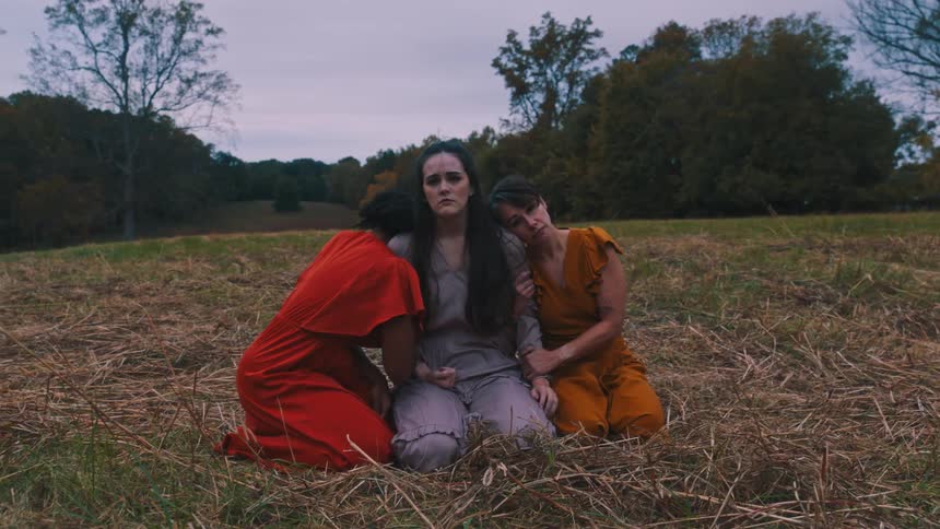 Three women in different colored dresses kneeling in a field performing a melancholic performance