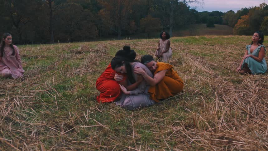 Group of women stands in a field of dry grass under a cloudy sky with some of them sitting around and others hugging each other in the center