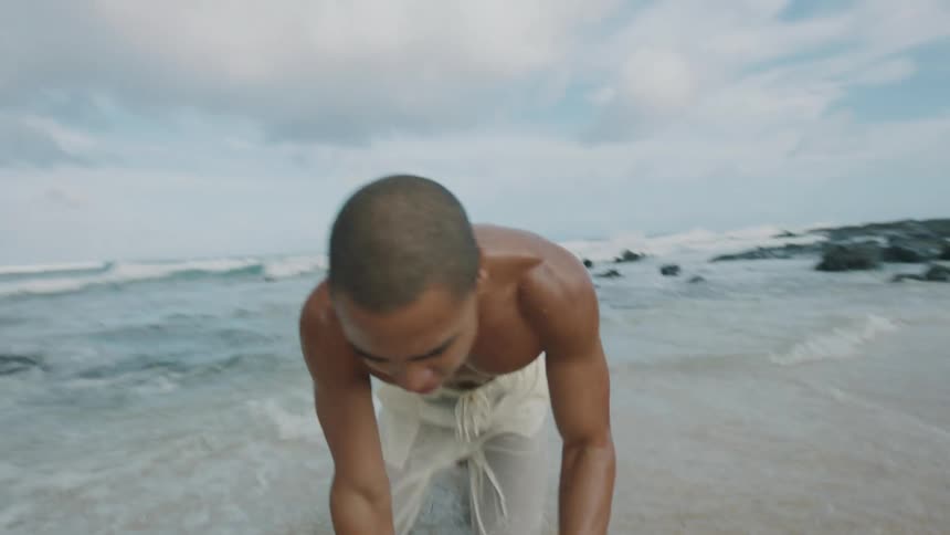 A muscular shirtless young man in white pants emerges from the ocean and collapses tired onto the sandy beach