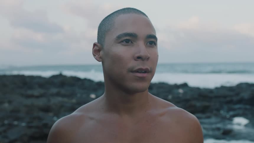 A bald muscular man stands shirtless on a rocky coastline looking away