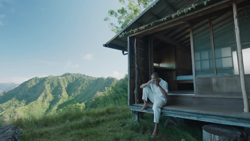 Man is sitting on the steps of a wooden cabin sipping from a mug and green hills in the background