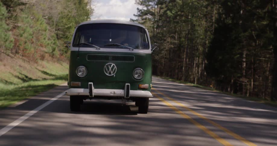 An old vw bus driving down a road in a forested area