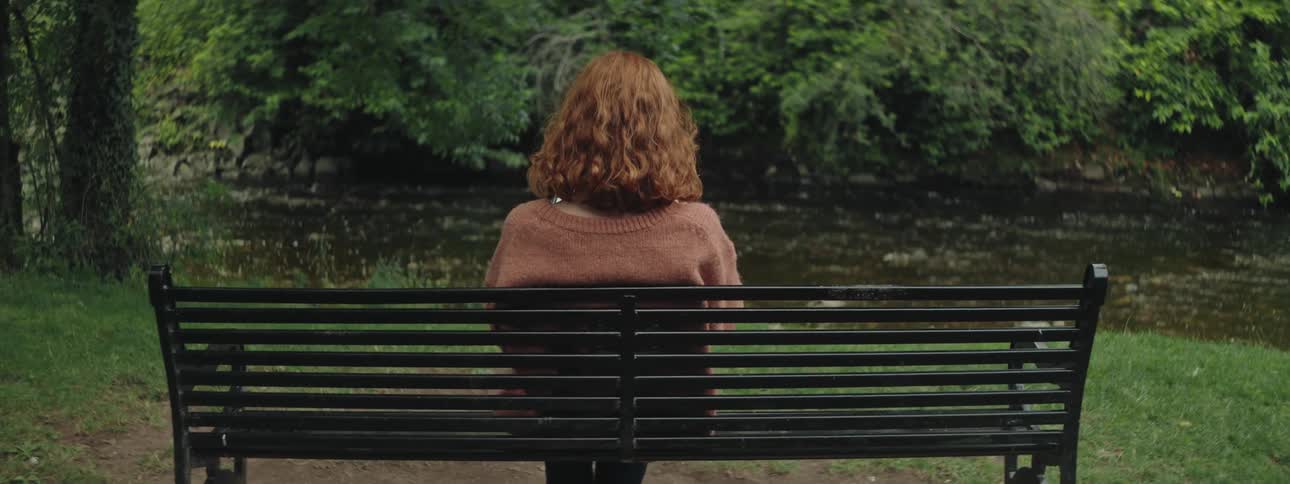 Back view of a redheaded woman sitting on a bench facing the river