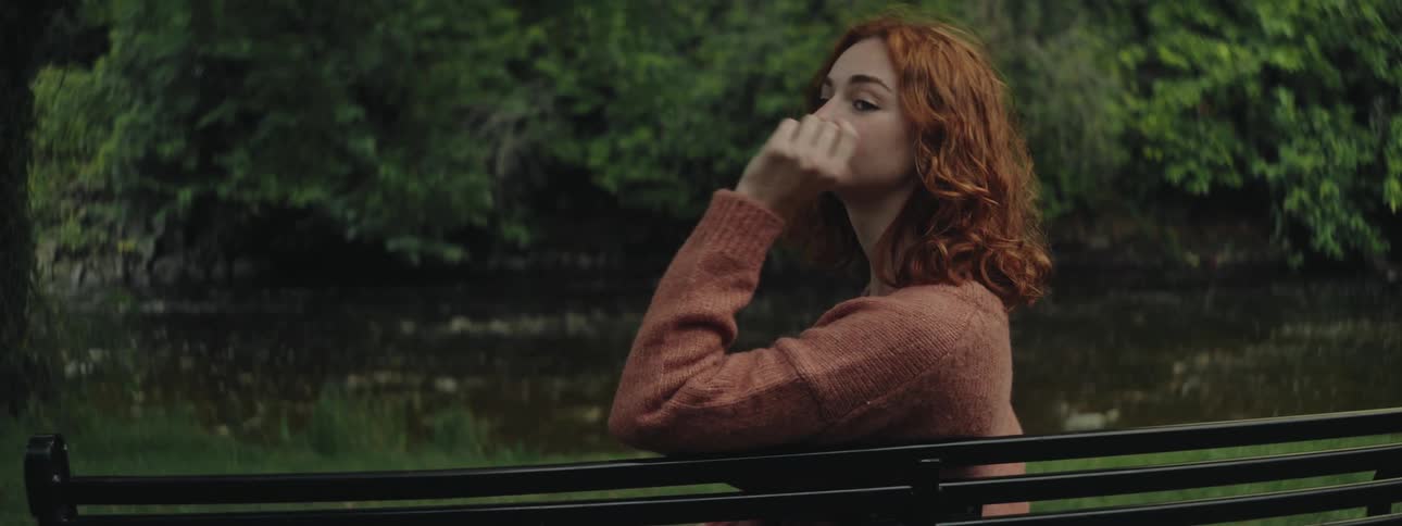 A young red headed girl sits on a bench by a river and turns her head to look behind her