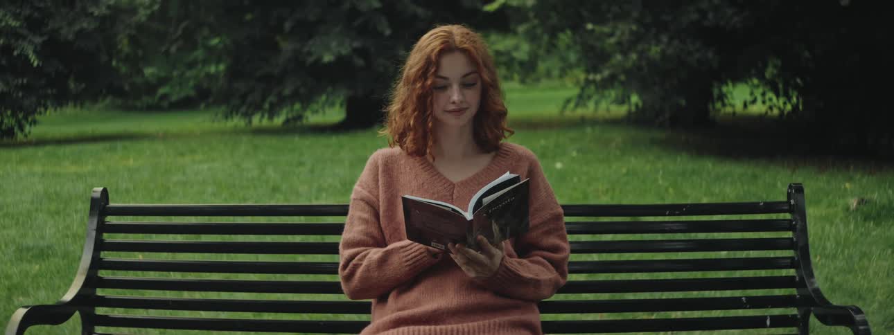 Redheaded woman smiles while reading a book on a park bench