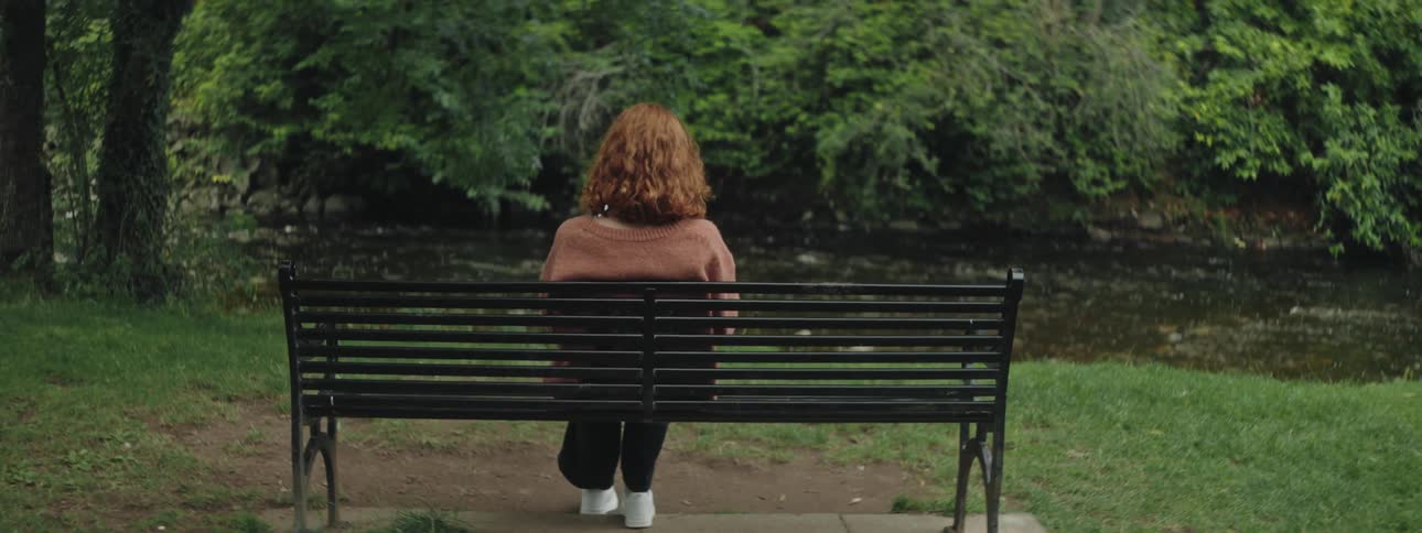A redheaded woman sits on a bench facing a river