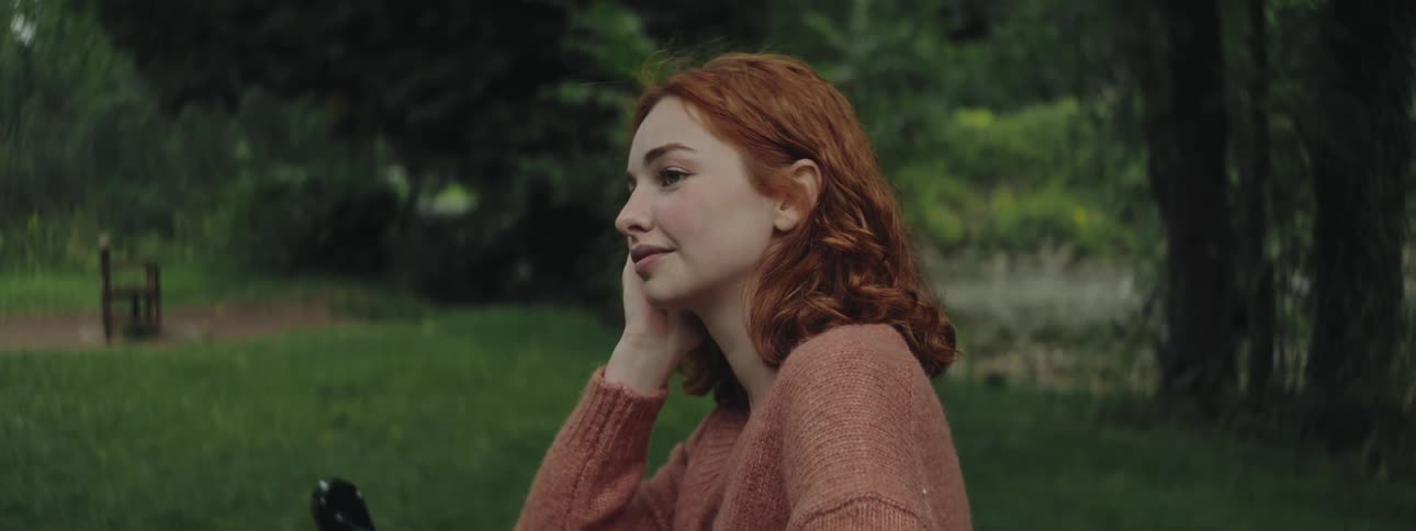 Red haired woman sits in a field near the woods and a water feature