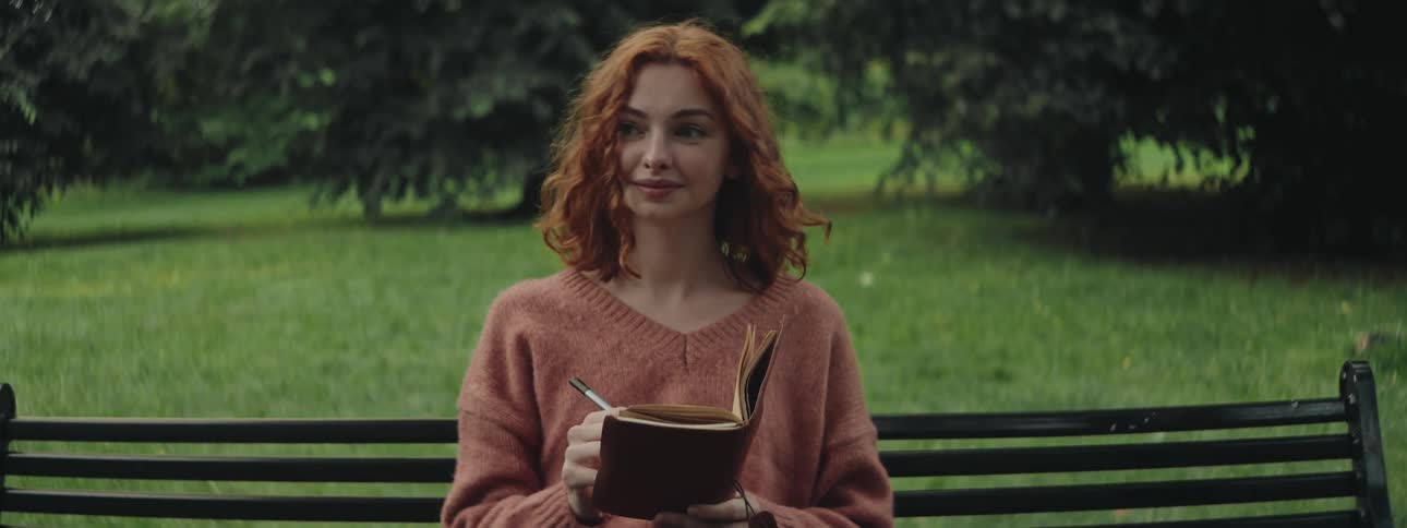 A young redhead woman in a pink sweater writes in a notebook on a park bench