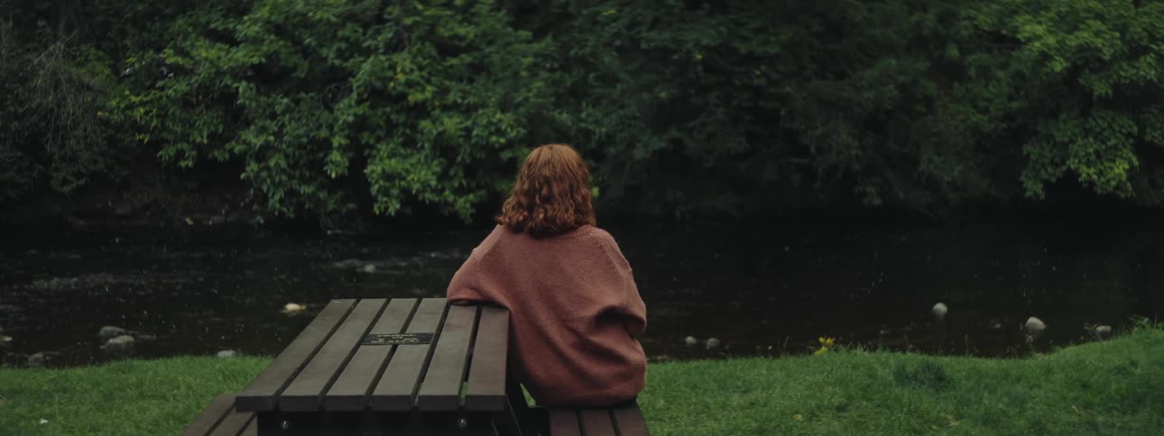 Redhaired woman sitting at a table in a square on the grass and facing a river