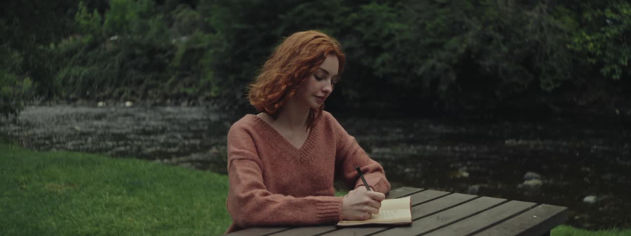 Thoughtful woman writing in a notepad while sitting on a bench by a stream