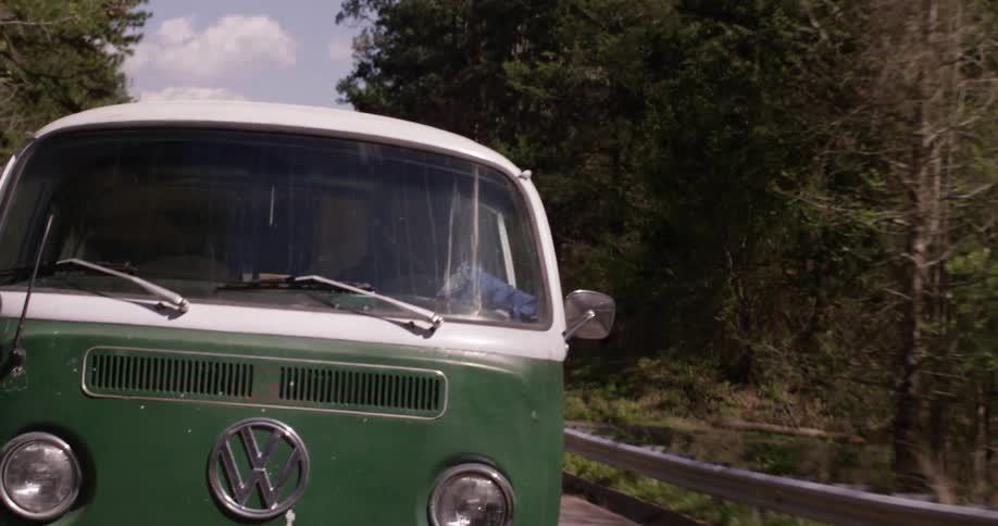 An old vw bus drives down the road through a forest with puffy clouds overhead