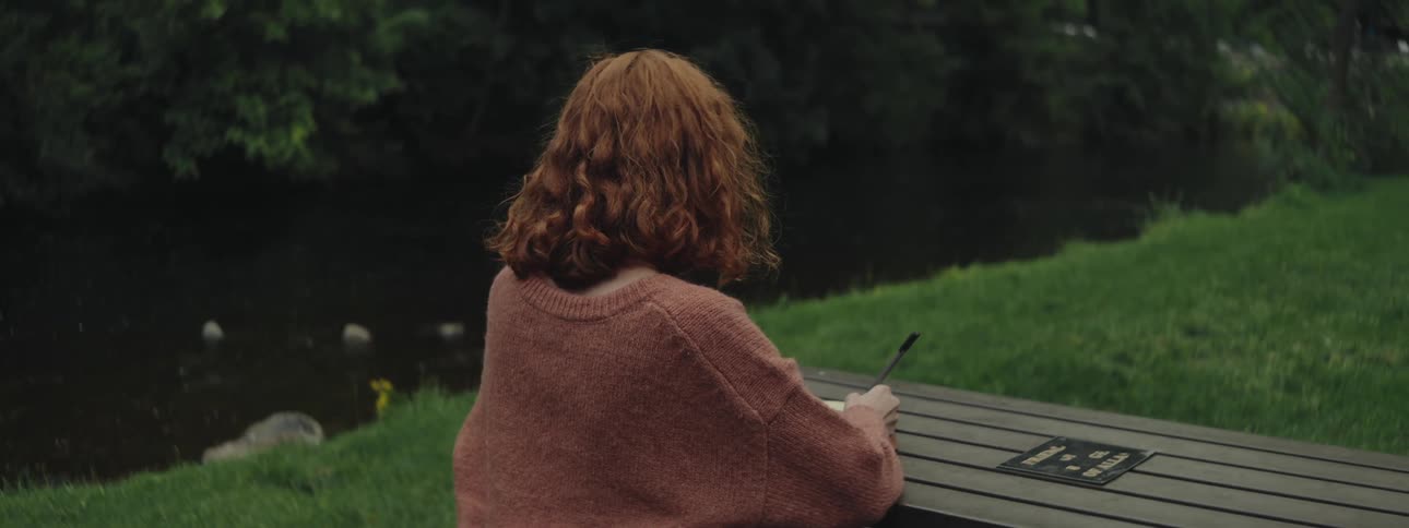 A redheaded woman sits a table by a river and writes in a book with a pen