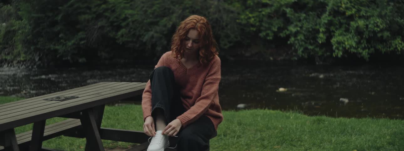 A teenage girl sitting at a picnic table near a river starts to tie her white shoe