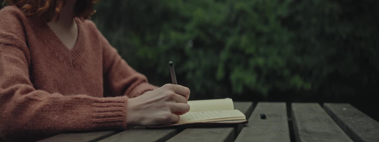 Woman in a sweater writes in a notebook on a wooden table with greenery in the background