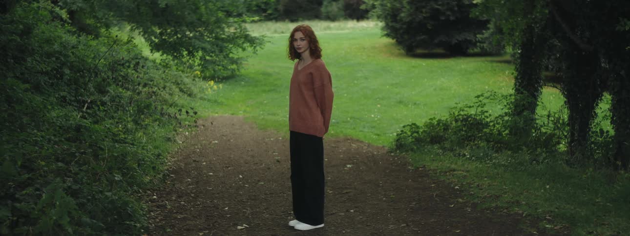A young redhead woman stands in a park looking at the camera