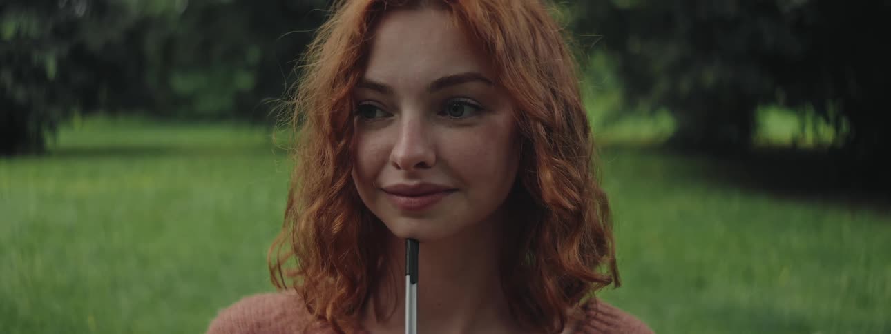 Portrait of a redhaired woman holding a pen while standing in an environment with grass and trees in the background