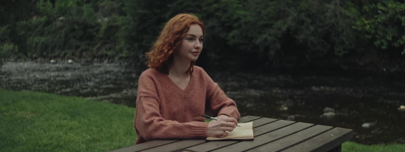 Redhaired woman sitting at a table writing in a notebook by a stream