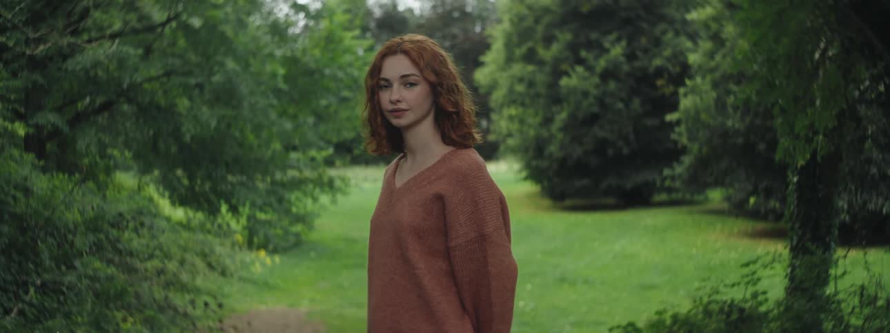 Redheaded woman smiles while posing outdoors in a landscaped grove