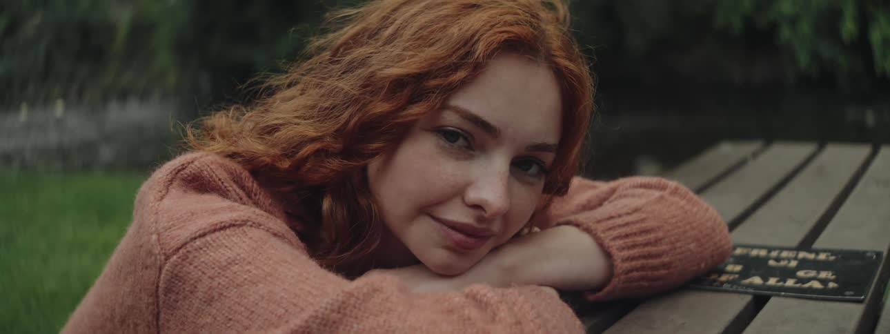 Portrait of a young redheaded woman sitting with her hands resting on a table in an outdoor space with a stream in the background