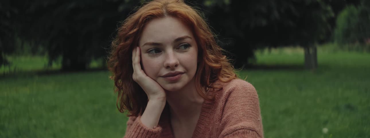 A teenage girl with red hair wearing a sweater rests her head on her hand while on a grassy plain