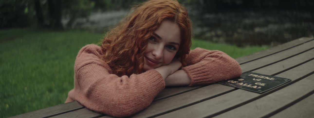 A teenage girl rests her head on her crossed arms on a picnic table by a river