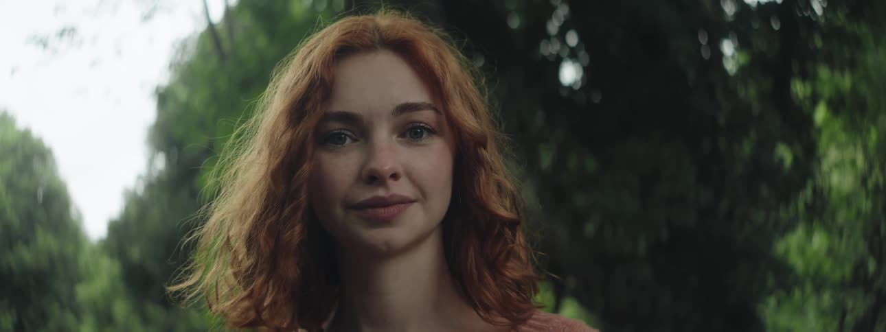 A young redhead woman stands in a park looking at the camera