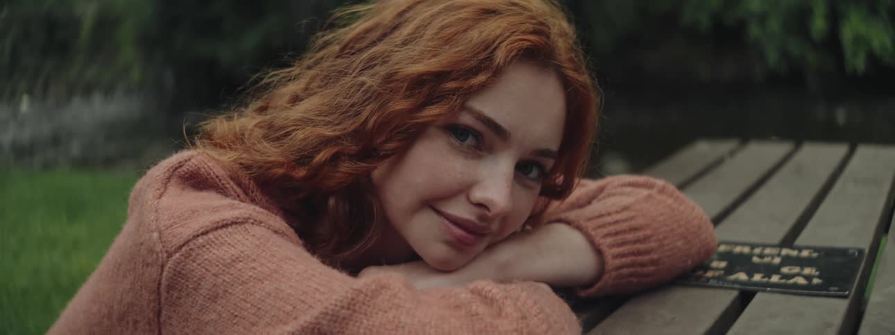 Smiling redhead rests her head on her crossed arms at a picnic table in the park during the daytime