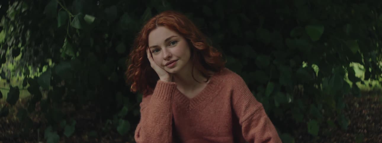 A young woman with wavy red hair wearing a cozy brown sweater resting her head on her hand and smiling gently set against a dark leafy background