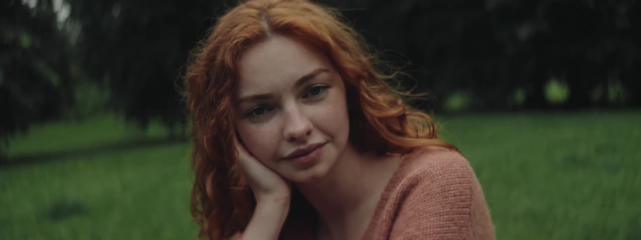 A young woman with red wavy hair wearing a soft brown sweater resting her head on her hand with a peaceful expression surrounded by a blurred green outdoor background