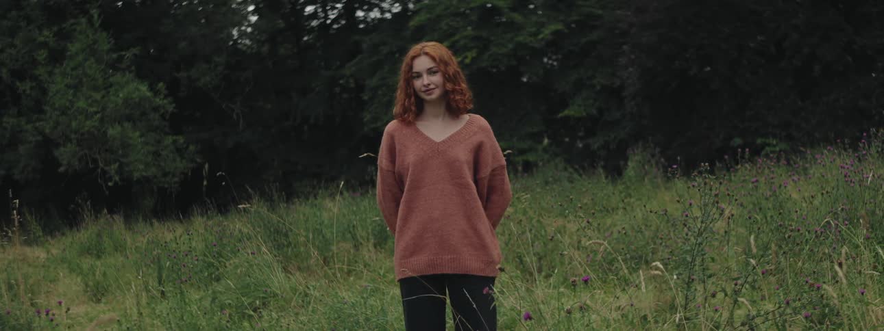 Redhead woman with her arms behind her back in a field with grass and trees in the background