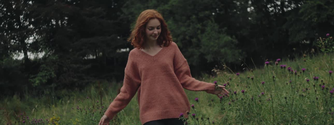 A woman with red hair and a brown sweater walks through a field of tall grass