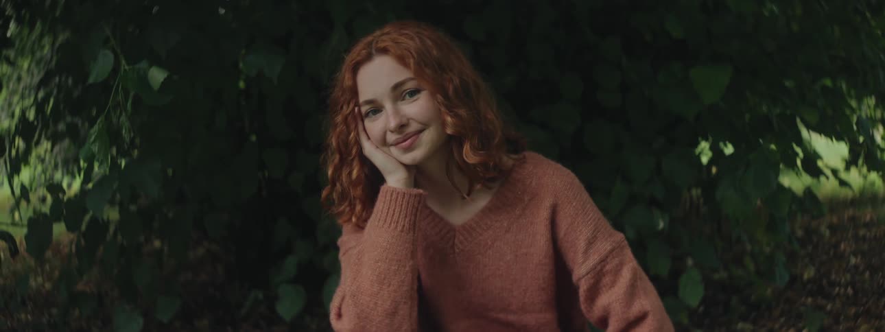 A teenager with red hair smiles while sitting under a tree