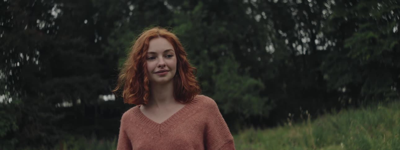 A red haired teenage girl smiles as she walks through a weedy grassland