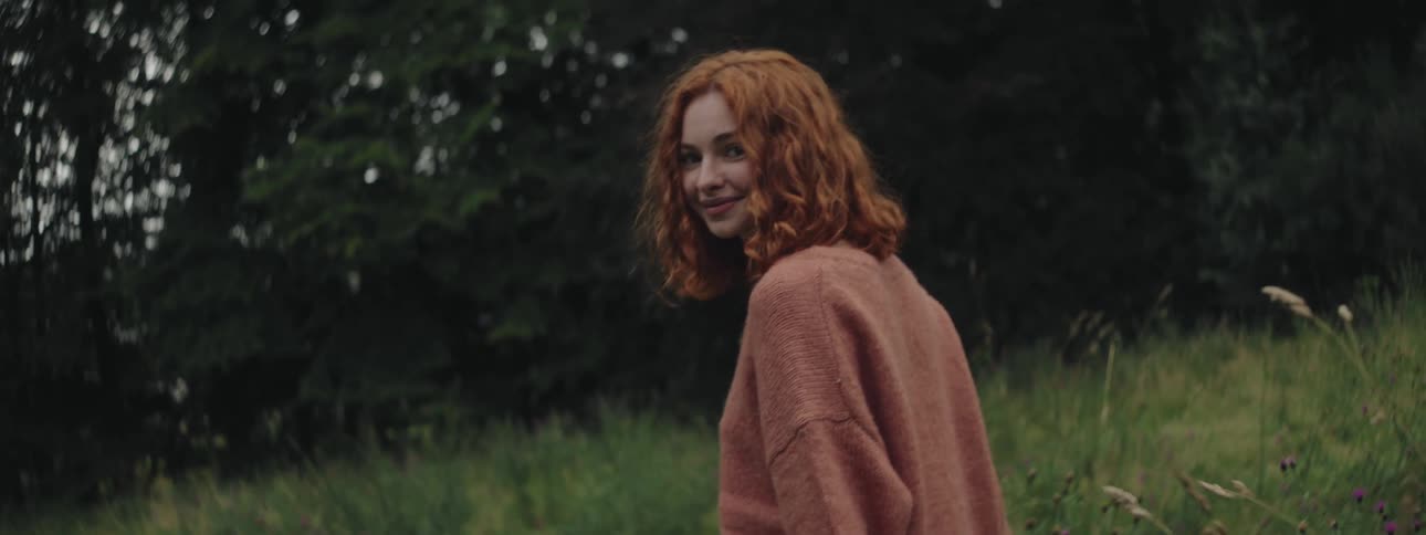 A woman with red wavy hair wearing a soft brown sweater smiles as she walks through a field of tall grass and green trees