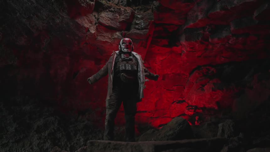 Individual wearing a red mask stands in a cavern lit by red colored lighting