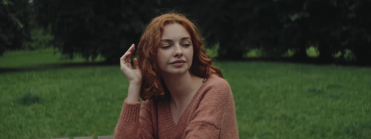 Redheaded woman with her hand on the back of her head sitting in an area with grass and trees
