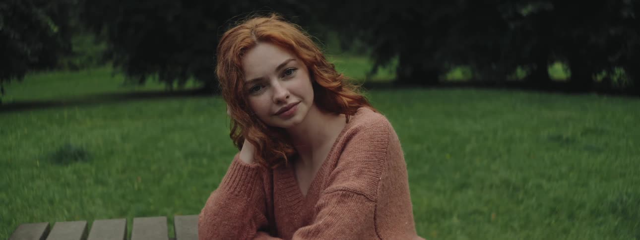 A red haired woman poses for a portrait outside while sitting at a bench