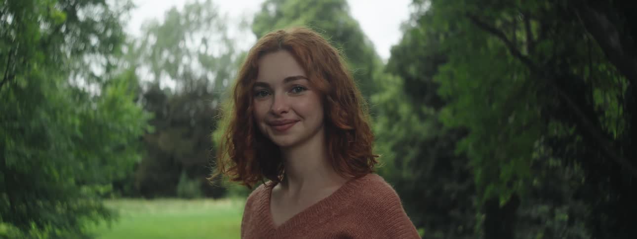 A red haired woman in a brown sweater smiles happily in a green park