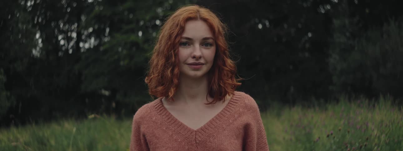 Redhaired woman in a longsleeved blouse with a happy facial expression in a field with trees in the background