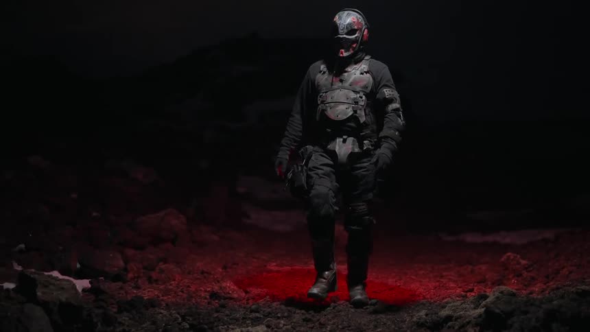 Person wearing armor and a mask walks over rough terrain lit by a red light at night