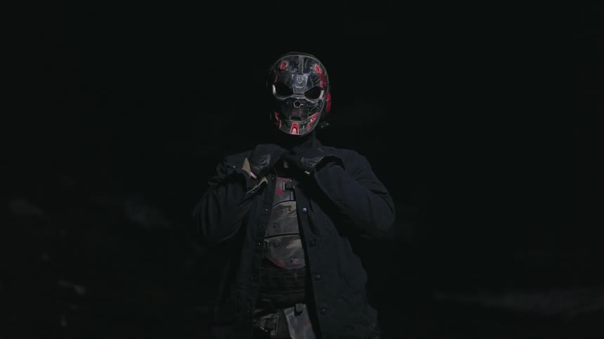 Person wearing a mask and armor stands among boulders under a light in a dark area