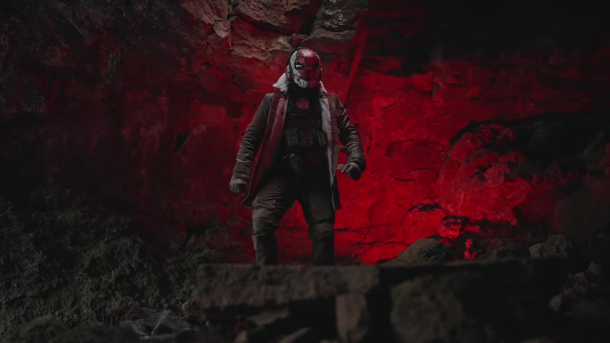 A masked figure wearing dark hooded jacket with a weathered appearance stands on a cave