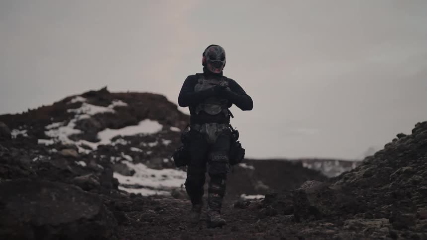 A masked figure in tactical gear walks along a snowy mountain ridge