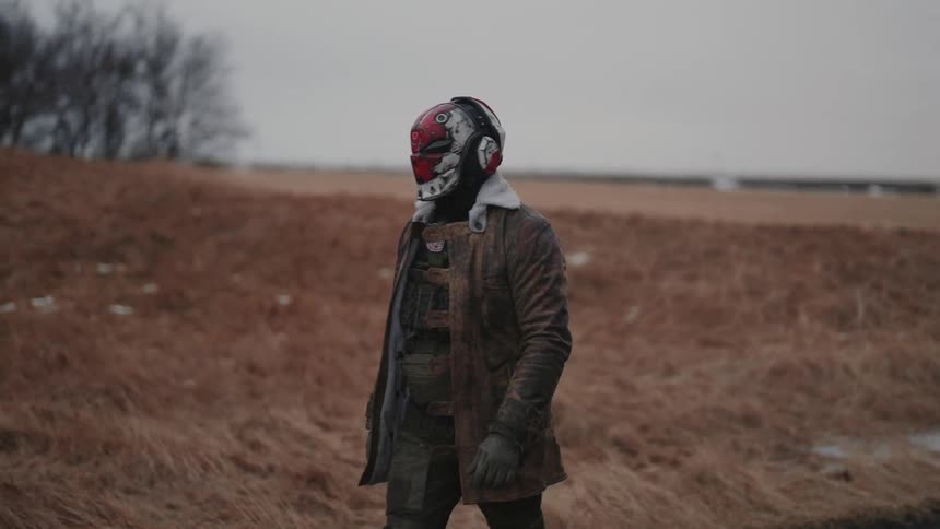 Person wearing a red and white mask with a jacket and vest walking through a field on a cloudy day