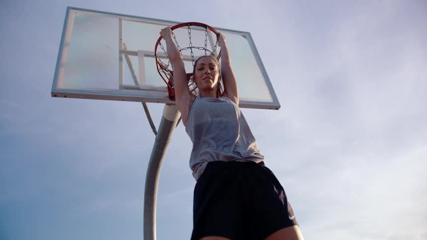 A female basketball player hanging from a basketball hoop and smiling