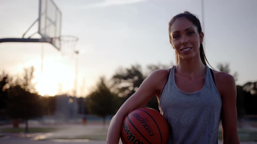 A woman standing on an outdoor basketball court holding a basketball with a hoop in the background during dawn or dusk
