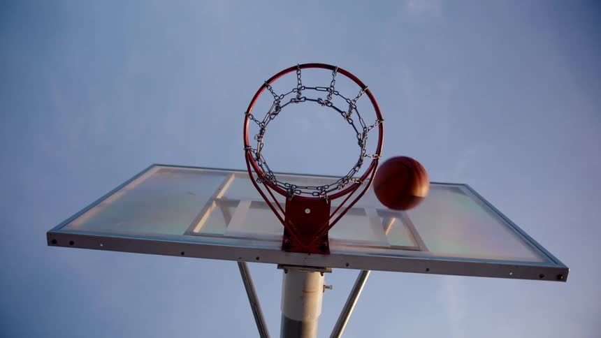 A basketball going through a hoop from below with a red and white net and a clear backboard against a light blue sky