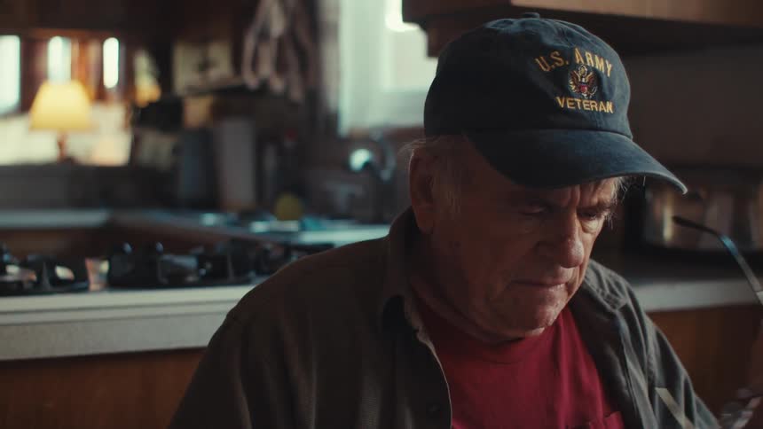 An old army veteran sits in a kitchen and thinks as he removes his glasses